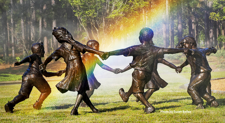 Bronze sculpture of children holding hands in a circle, set in a wooded garden with sunlight creating a rainbow effect through mist in the background.
