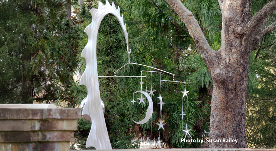 White sculptural form resembling a dragon’s tail supports a hanging mobile of crescent moon and star shapes, displayed outdoors among trees.