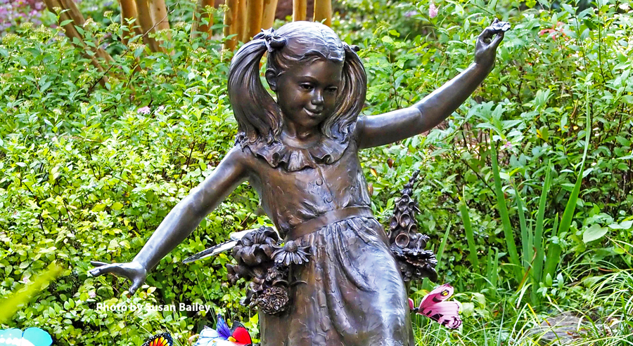 Bronze sculpture of a young girl balancing with outstretched arms in a garden, surrounded by flowers, greenery, and decorative butterflies.