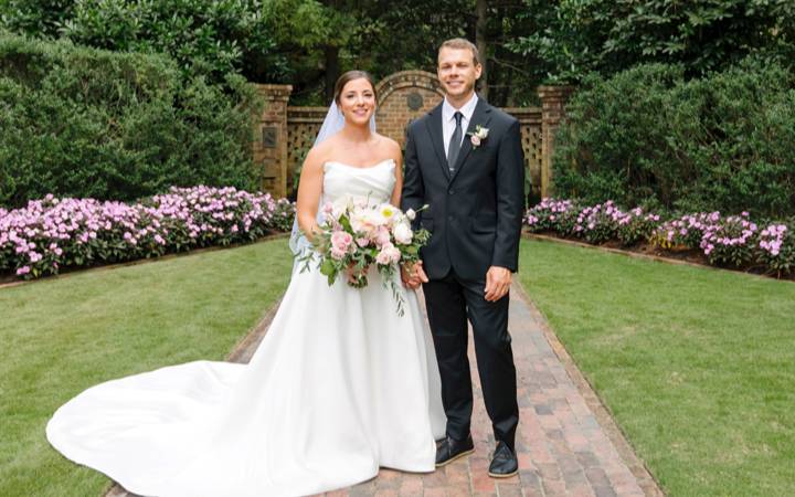 Bride and groom stand together on a brick pathway in a landscaped garden courtyard, surrounded by greenery and flowering plants.