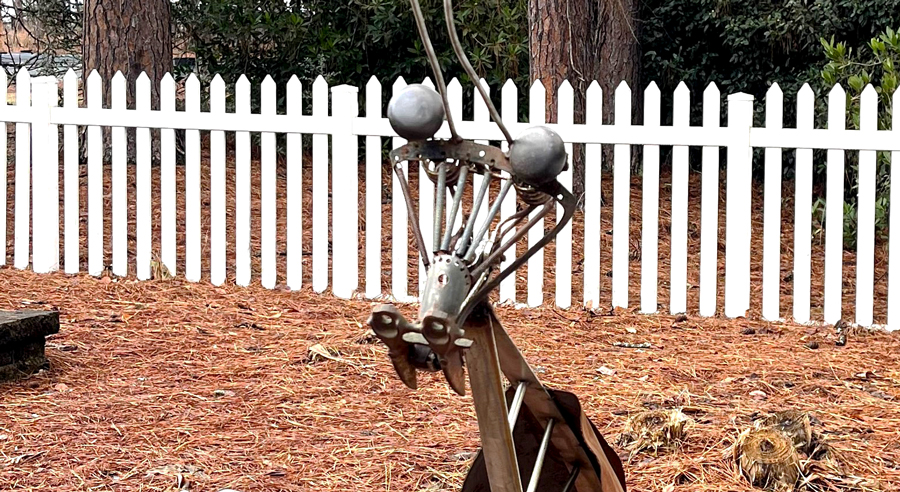 Tall metal insect sculpture made from industrial parts, featuring long antennae and spherical eyes, standing along a mulched garden path bordered by a white picket fence and pine trees.