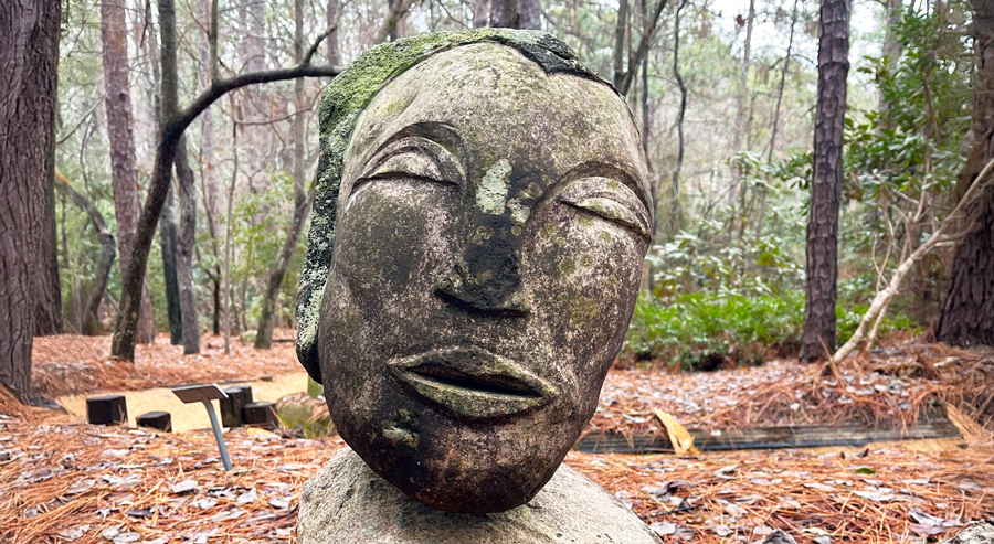 Stone sculpture of a serene human face with closed eyes, weathered with moss and lichen, resting atop a natural rock base among pine needles and trees in a wooded garden setting.