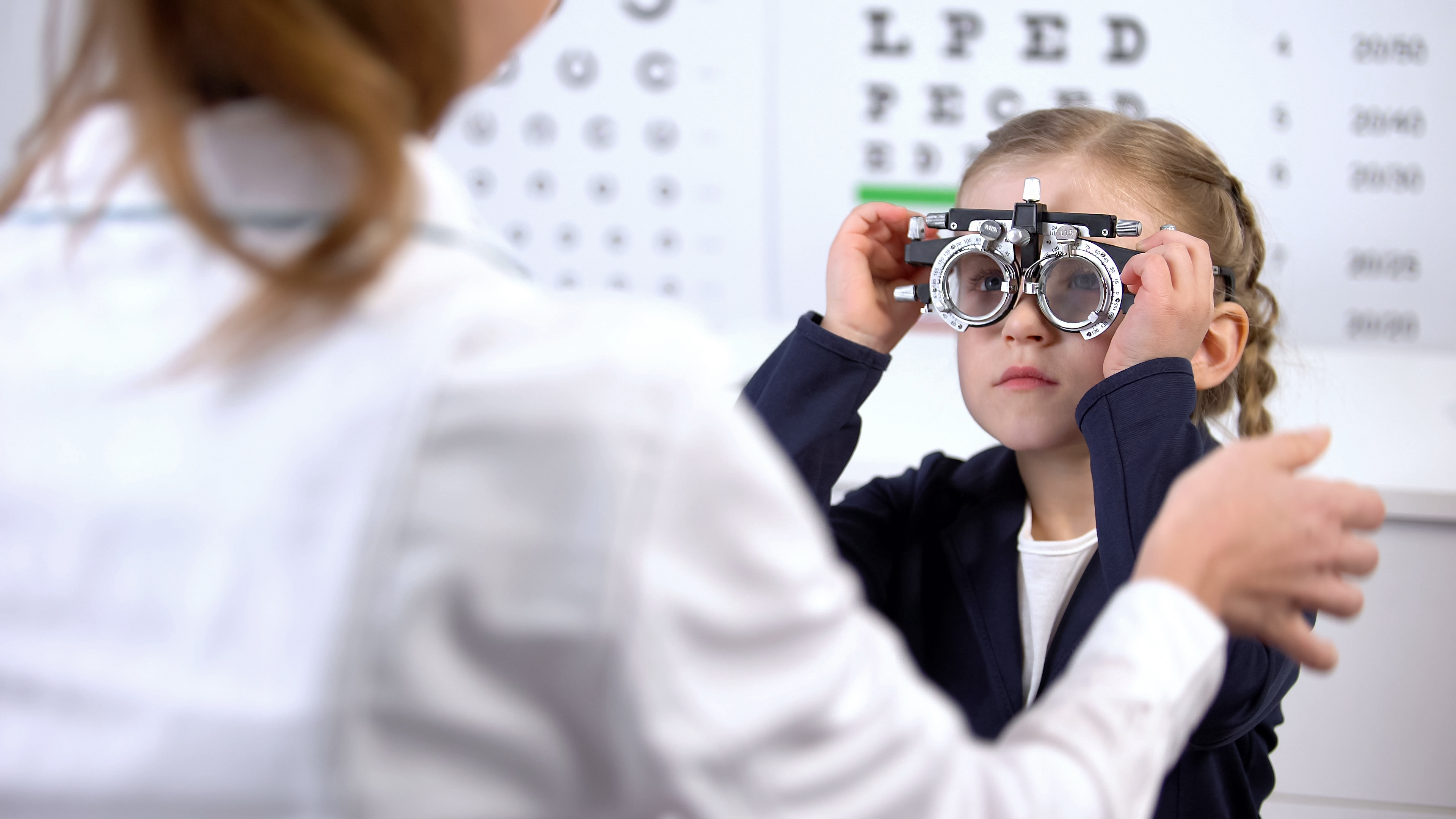 A young girl sits in an eye clinic wearing a phoropter while an optometrist adjusts the lenses during a vision test. An eye chart is visible in the background.