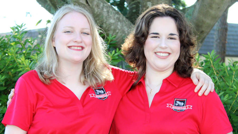Two female Sandhills Community College student ambassadors wearing red polo shirts stand outdoors with arms around each other, smiling in front of greenery.