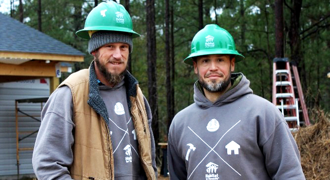 Two Habitat for Humanity construction team members wearing green hard hats and work clothing stand outdoors at an active build site with tools and equipment visible in the background.