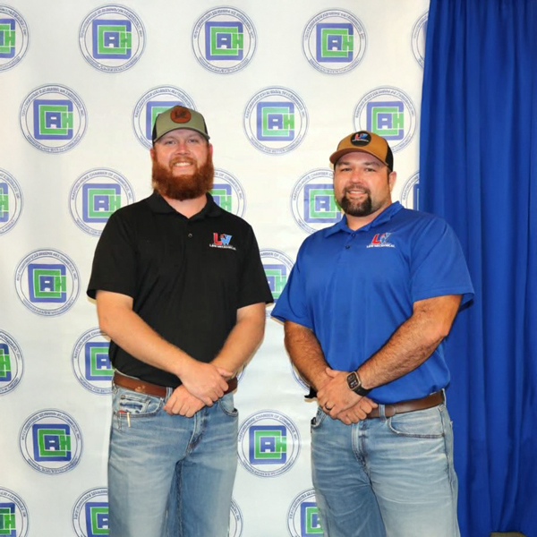Tyler Lewis and Chris Wright of L&W Mechanical LLC stand side by side in branded polos and caps in front of a Raeford-Hoke Chamber backdrop at a business recognition event.