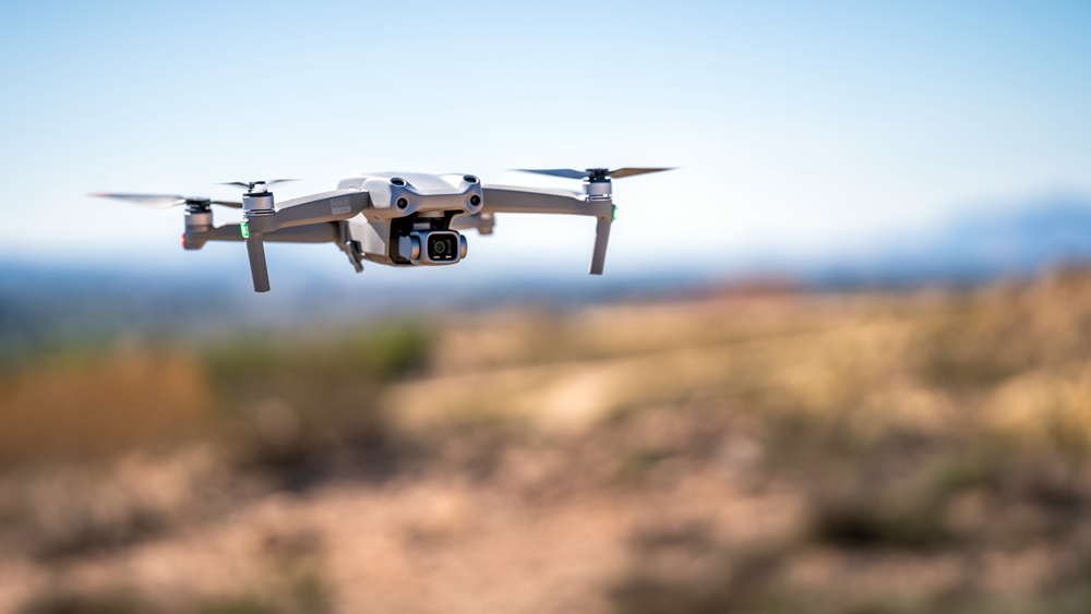 A drone hovers in flight above an outdoor landscape with its camera facing forward.