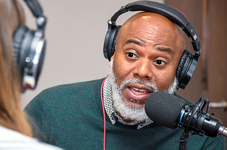 A podcast host wearing headphones speak into microphones while recording in a studio.