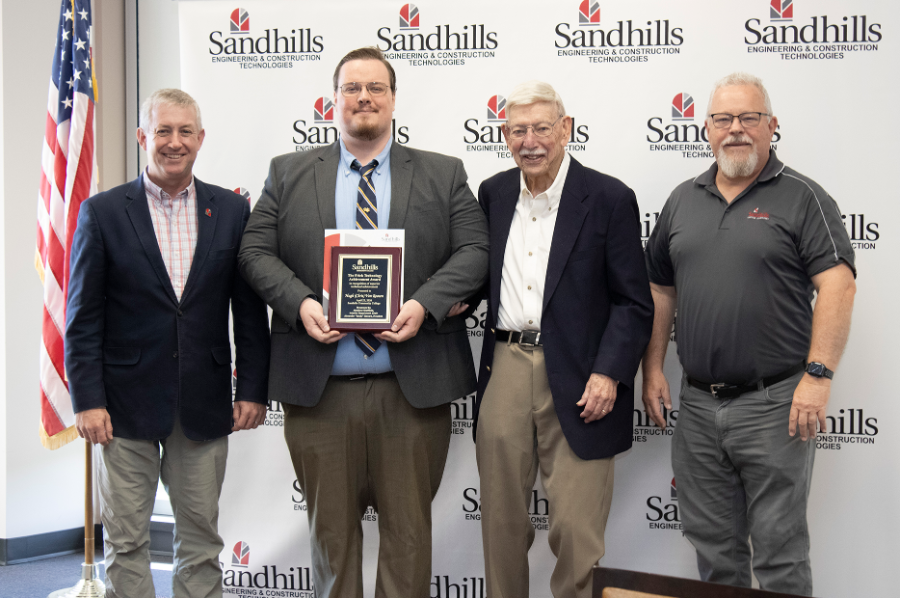 Four men stand in front of a Sandhills backdrop as award recipient Chris Van Roosen holds a plaque during the Frick Technology Achievement Award presentation.