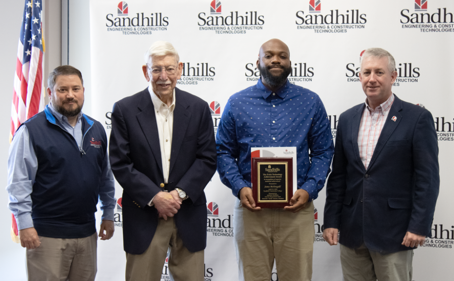 Four men stand in front of a Sandhills backdrop as award recipient James McDougald holds a plaque during the Frick Technology Achievement Award presentation.