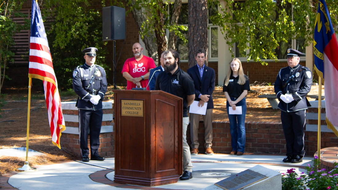 Timothy Justice speaks at a podium during an outdoor ceremony at Sandhills Community College, flanked by uniformed color guard members.
