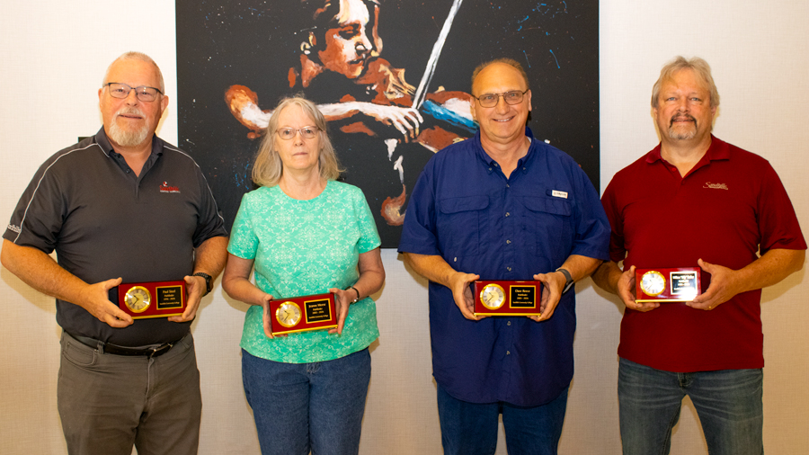 Four Sandhills Community College employees standing with service awards during a recognition ceremony in front of a violin artwork display.